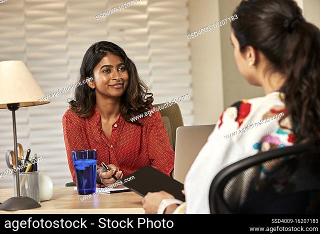 portrait of young ethnic woman sitting at desk with laptop computer talking to co-worker sitting across desk