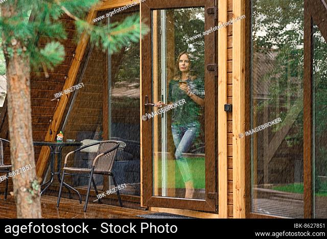 Young woman with a cup of herbal tea stands on the doorstep of a forest cabin and looks at the rain outside
