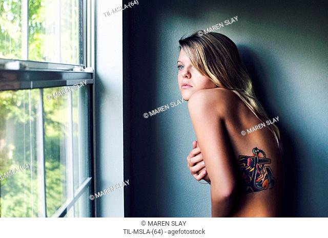 Close up of naked young woman with blonde hair standing alone in a room looking out of window
