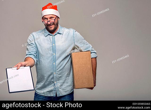 Cheerful attractive young man in a Santa Hat carrying or delivering a large brown cardboard box for Christmas, over grey with copy space