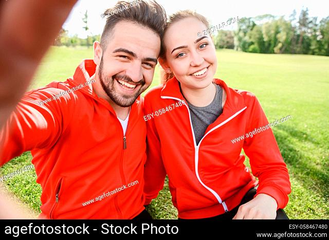 Closeup portrait of sport man and woman making selfies in green park or forest while sitting on green grass and resting after jogging