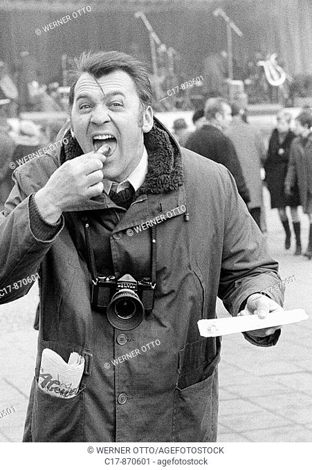 Sixties, black and white photo, people, humour, man with a camera and wide opened mouth bites into a frying sausage and makes faces, aged 30 to 35 years