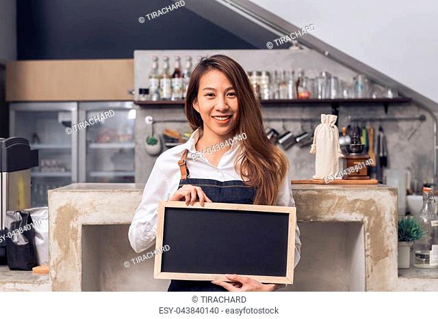 Young Asian female barista in denim apron hold a chalkboard with a beautiful smile in her own coffee shop welcome her customer to try a new coffee menu