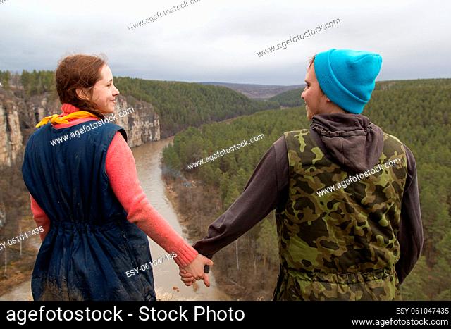 Rear view of couple hikers standing on the edge of a cliff over the mountain river and holds hands each other, telephoto shot