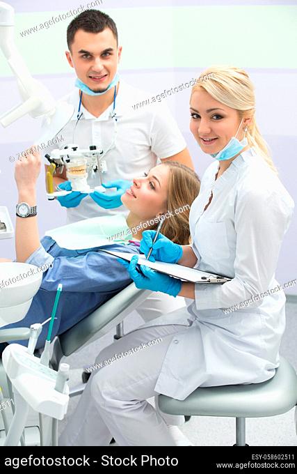 Attractive girl in blue shirt and patient bib on the patient chair in the dental cabinet. She holds a mirror in the left hand