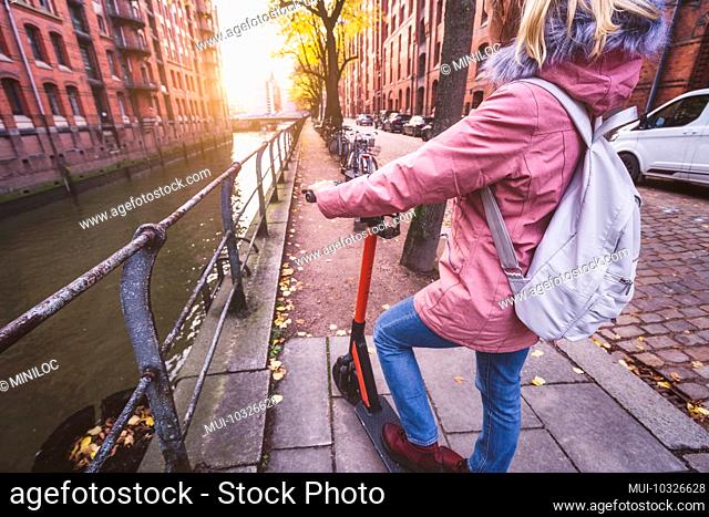 Rear view of a woman with a backpack and e-scooter, view of the historic warehouse district in Hamburg in the warm light of the sunset, Germany, Europe