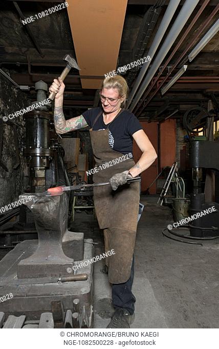 female blacksmith working at ambos with hammer on redhot steel, steamhammer in the background