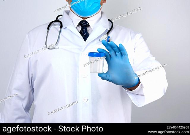 male doctor in a white coat and tie stands and holds a empty plastic container for urine specimen, wearing blue sterile medical gloves