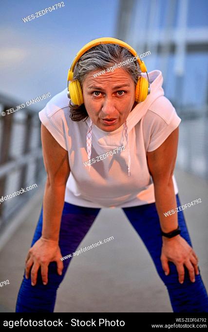 Mature woman wearing wireless headphones taking break from run at bridge