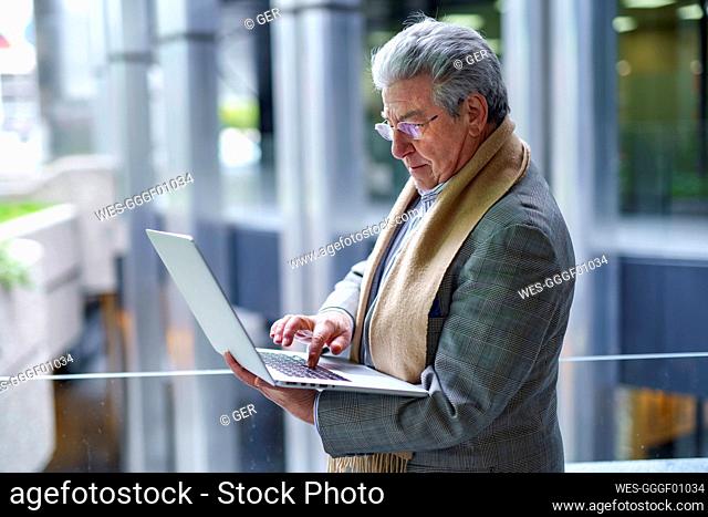 Senior businessman working on laptop standing by glass railing
