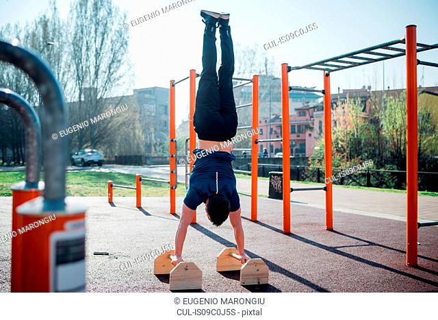 Calisthenics at outdoor gym, young man doing hand stand on exercise equipment, rear view