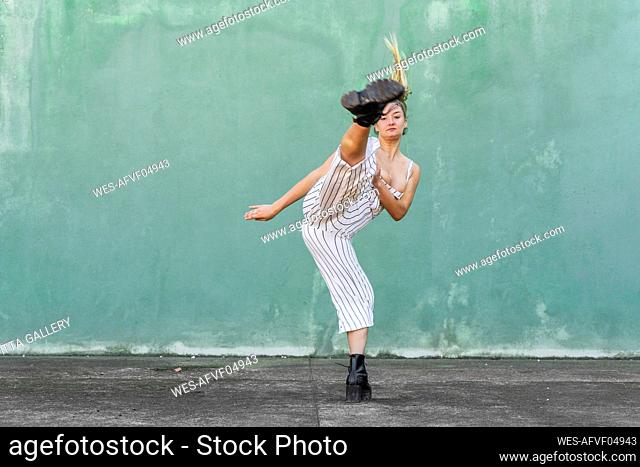 Young woman doing a high kick in front of green wall