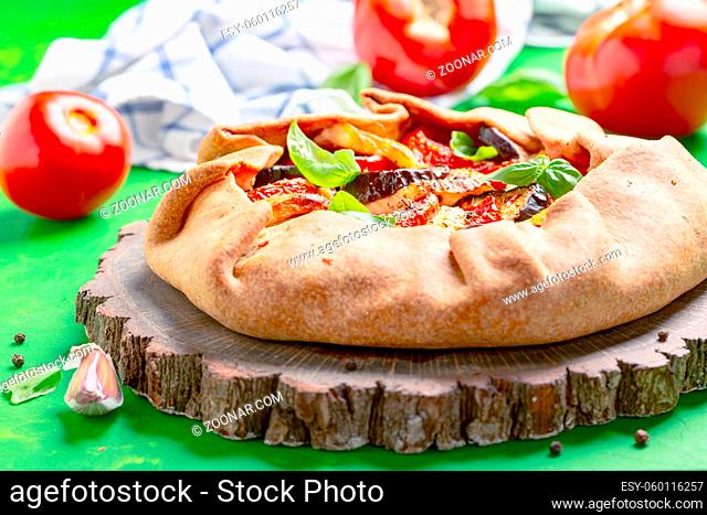 Open rye pie (galette) with tomatoes, eggplant, mozzarella and basil is served on a wooden serving board. Concept of baking homemade vegetable pies