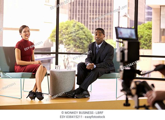 Black man and white woman on TV interview set look to camera