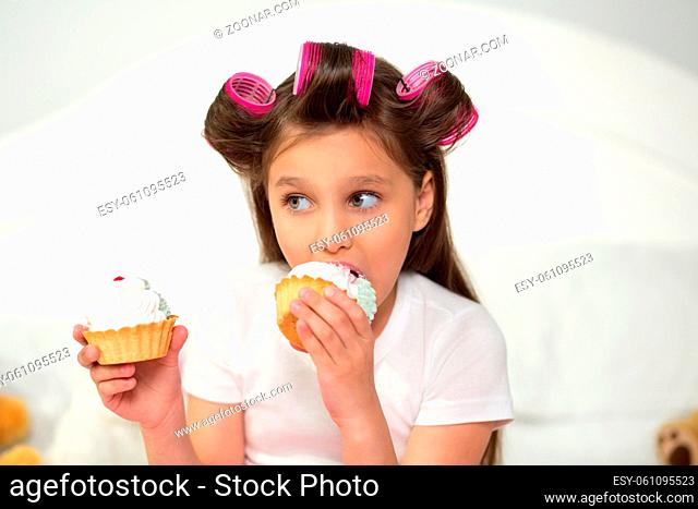 Charming young girl holding cupcake in each hand. Lovely little girl with hair curlers holding two cupcakes close portrait