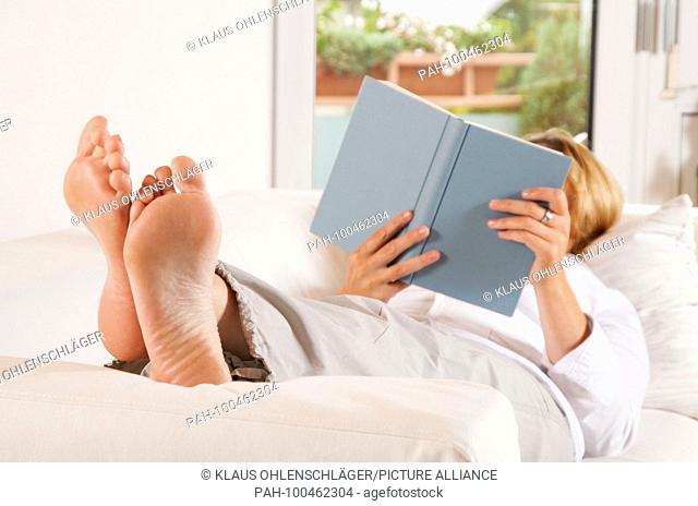 Woman is lying on the sofa, has her feet up and is reading a book. | usage worldwide