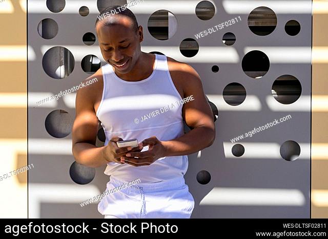 Smiling man text messaging through smart phone standing in front of wall
