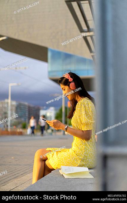 Young woman using smart phone and listening music while sitting on bench in city
