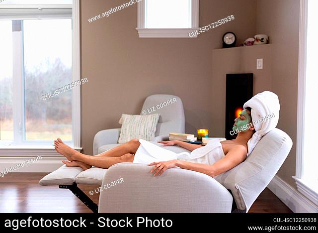 Woman with green face mask and head wrapped in towel relaxing in lounge chair