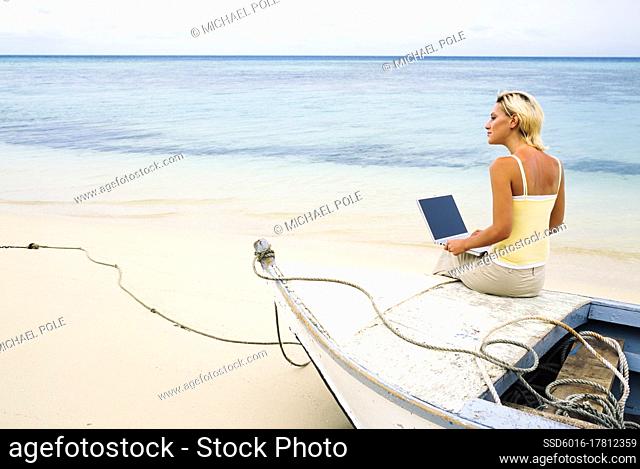 Young woman sitting on the bow of a small boat with her laptop computer on a tropical beach
