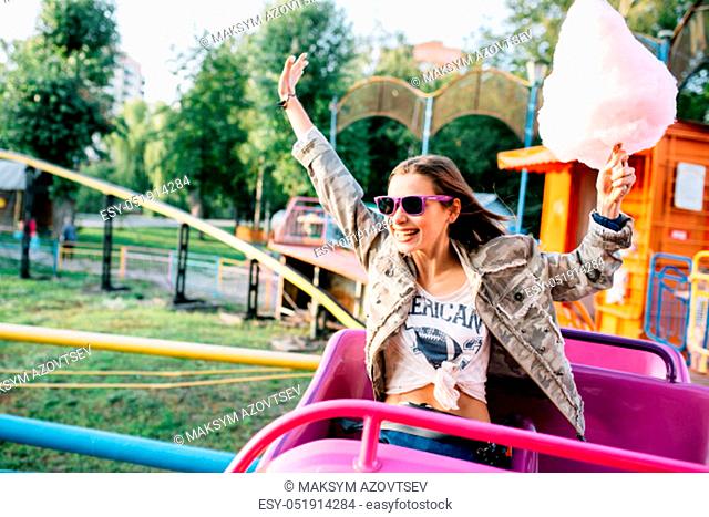Attractive cheerful girl in sunglasses riding a roller coaster, having fun, raised her hands up, holding a cotton candy. Dressed in stylish jacket