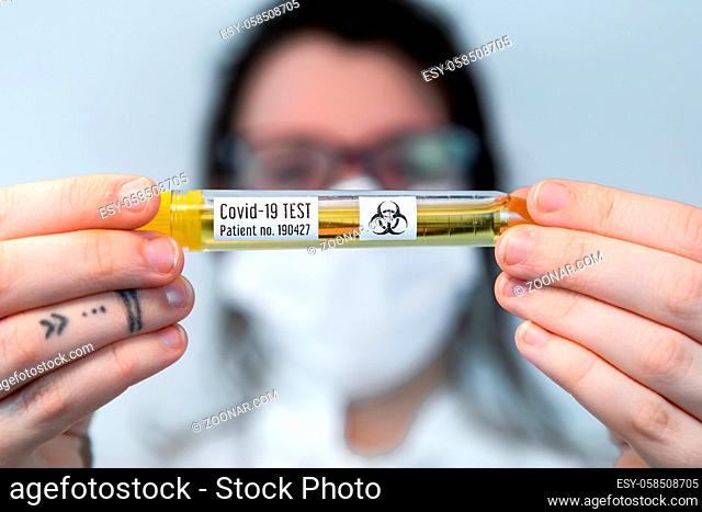 Selective focus closeup vial filled with yellow liquid labeled Covid-19 test held by tattooed hands of out of focus nurse over white background