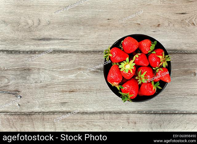 Tabletop view, small black ceramic bowl with strawberries, gray wood desk under. Space for text on left side