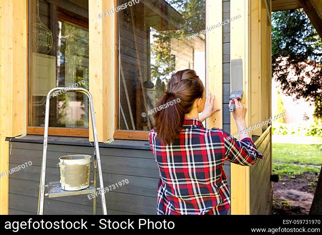 Woman worker painting wooden house exterior wall with paintbrush and wood protective color home improvement concept