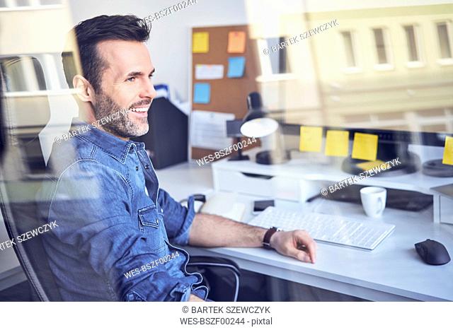 Smiling man looking through window in office sitting at desk