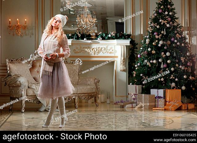 Christmas evening. Young beautiful blonde woman with cup of coffee in classic apartments a white fireplace, decorated tree