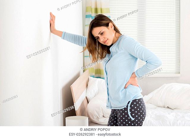 Woman Suffering From Back Ache Standing Against Wall