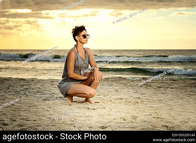 beautiful woman in a gray dress crouches on the sand at the beach at sunset
