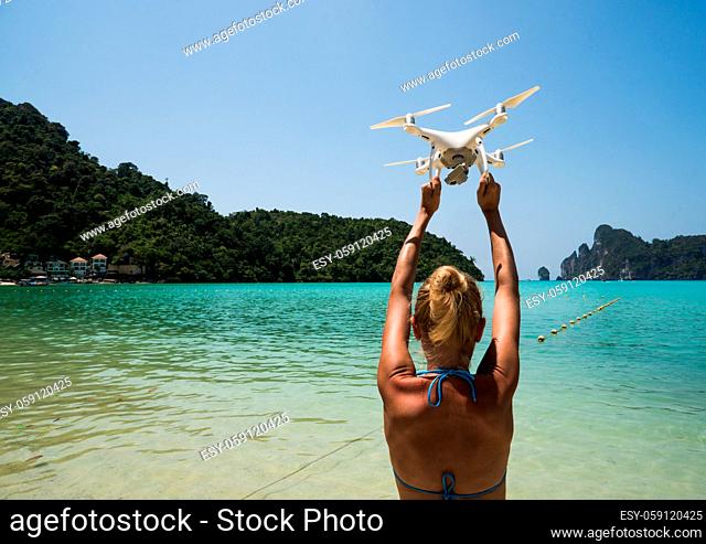 Back view of woman standing on the beach with raised hands holding drone over beautiful sea and summer blue sky background