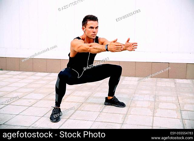 Full length of young concentrated sportsman doing squats during workout outdoors, listening to music in earphones. Wearing black sportswear