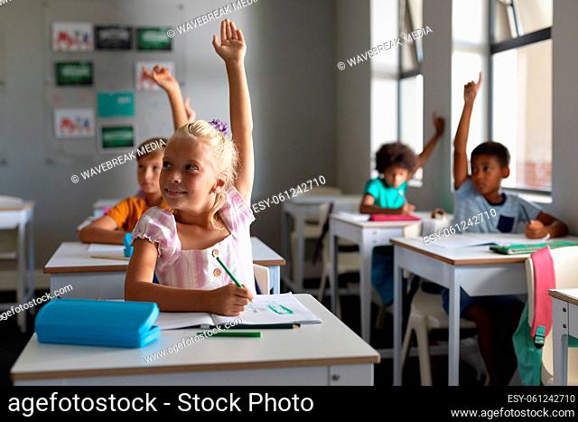 Multiracial elementary school students with hands raised sitting at desk in classroom
