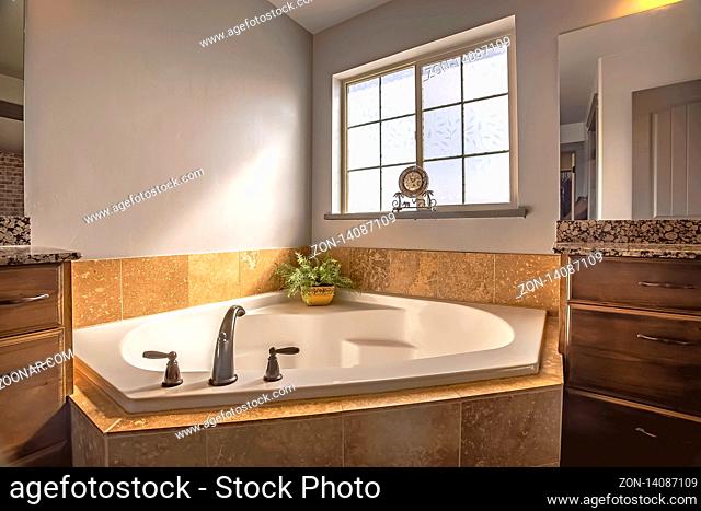 Built in bathtub at the corner of a bathroom with frosted glass window. The tub is between two vanity units with wooden cabinets and large mirros