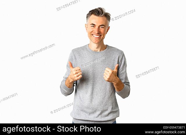 Portrait of handsome middle-aged guy with grey hairstyle, showing thumbs-up and smiling, approve something good, like idea, standing over white background
