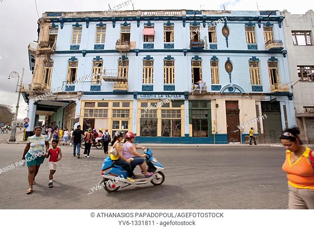 Typical Central Havanas Road With Scooters And People Crossing The Street While An Ex Luxurious Blue Building Is Noticeable At The Background, Centro Habana