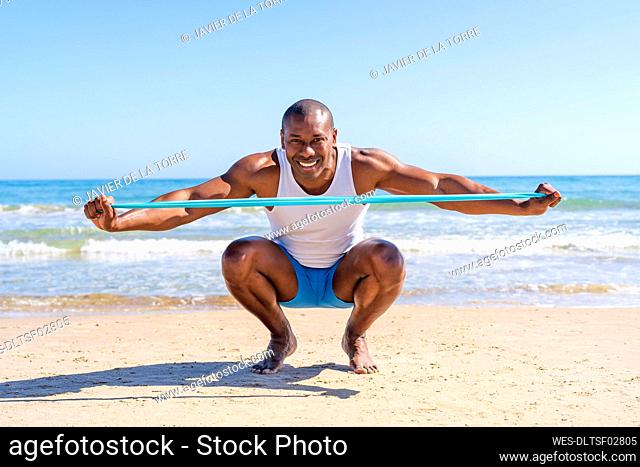 Smiling sportsperson with resistance band crouching at beach