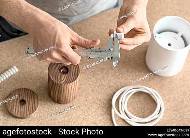 Process of measuring in the workshop. Man is using a caliper on the white metal detail. Under his hands there are wooden and metal cylindrical billets, cable