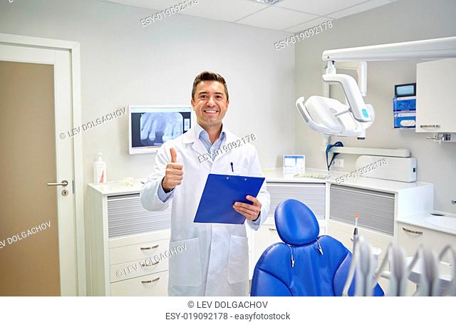 people, medicine, stomatology and healthcare concept - happy middle aged male dentist in white coat with clipboard showing thumbs up at dental clinic office