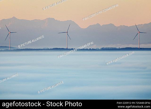12 September 2022, Baden-Wuerttemberg, Uttenweiler: Wind turbines are illuminated by the rising sun in the morning, while the Alps