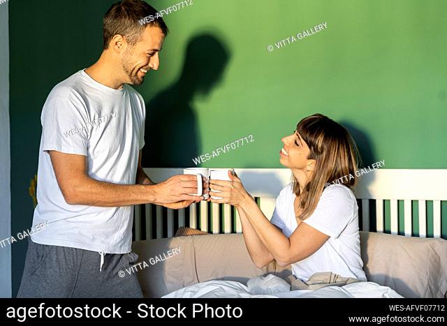 Smiling boyfriend and girlfriend toasting with coffee while sitting on bed at home