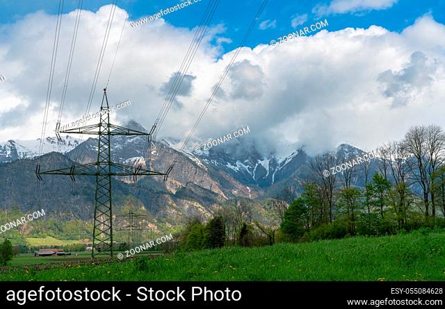 high voltage power line and lattice cross in agricultural farm fields in midst of an idyllic Swiss Alps mountain landscape