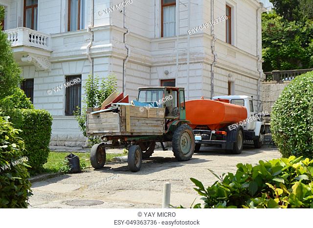 Old tractor with garbage in the back is near the watering machine, cleaning equipment for cleanliness in parks on a Sunny summer day