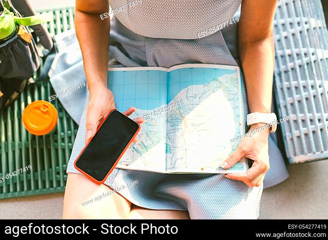 Theme travel planning. The top view hands Caucasian woman uses smart phone and studies the tourist map, passes route, navigates through the paper map