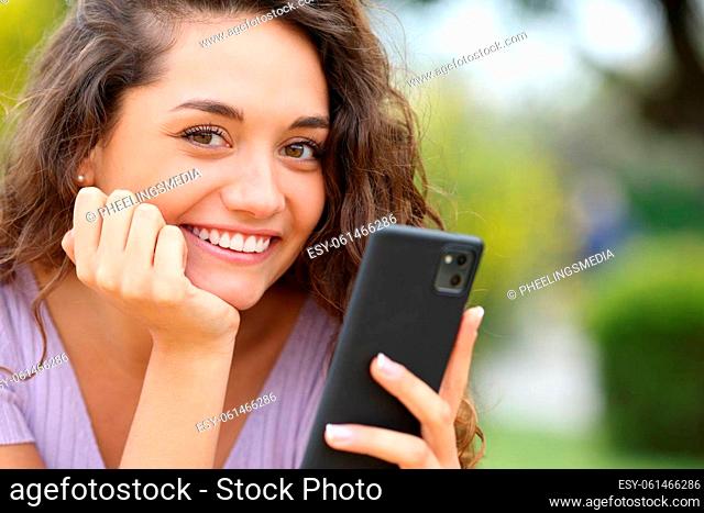 Happy beautiful woman holding cell phone looks at you in a park