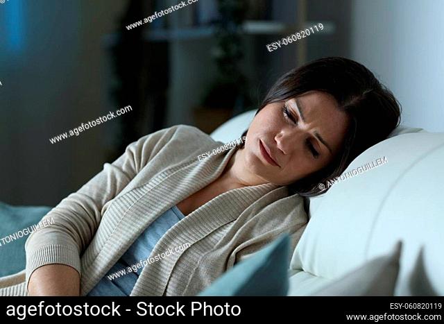 Sad woman looking down sitting on a couch in the living room at home in the night