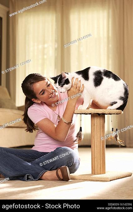 Teenaged caucasian girl sitting cross-legged on rug in living room petting her cat, cat sitting on top of scratching post