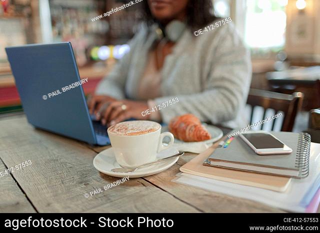 Woman working at laptop next to croissant and cappuccino in cafe